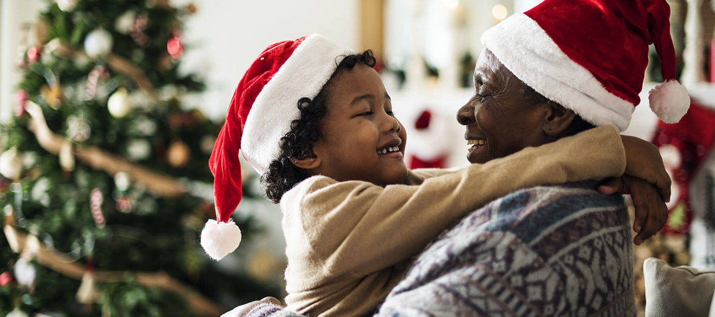 A grandfather holding his grandson in front of a Christmas tree, both wearing Christmas hats