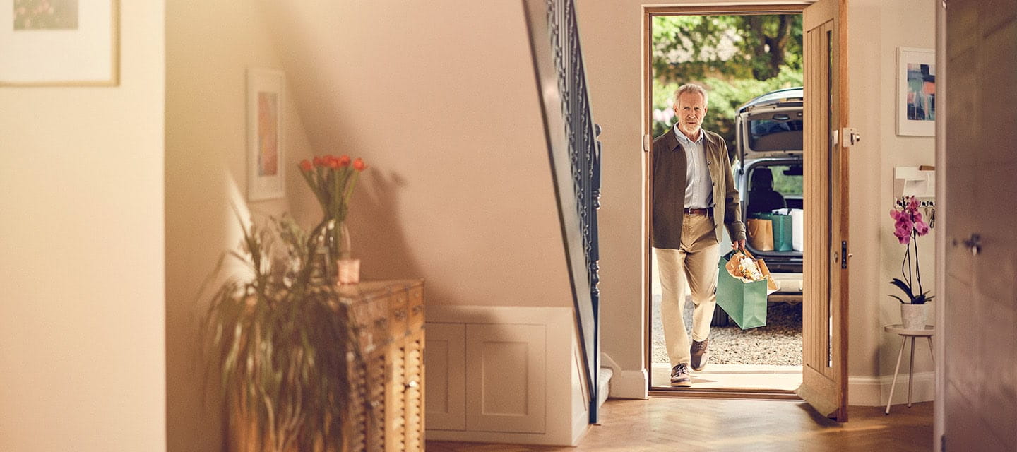 A mature man entering his home carrying a bag a shopping, you can see the boot of his car on the driveway containing more shopping.