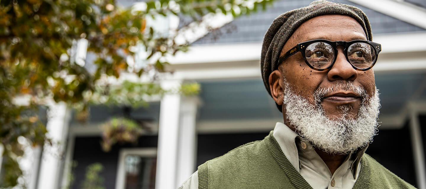 A stylish mature man with a grey beard standing in front of his house