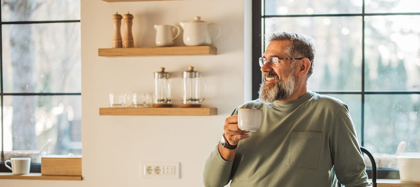 A mature man stood in his kitchen smiling while enjoying a cup of coffee