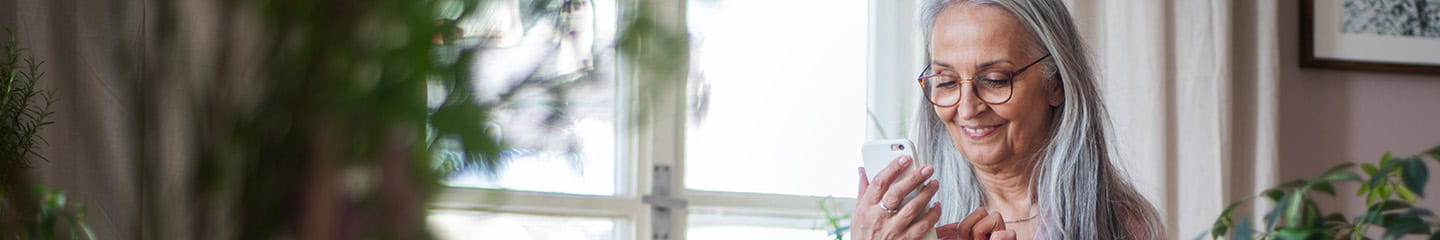 Fashionable senior woman standing by desk and text messaging at home