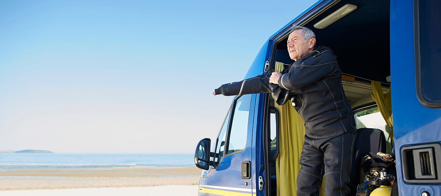 A senior diver puts on a wetsuit in his camper van parked beside the beach.