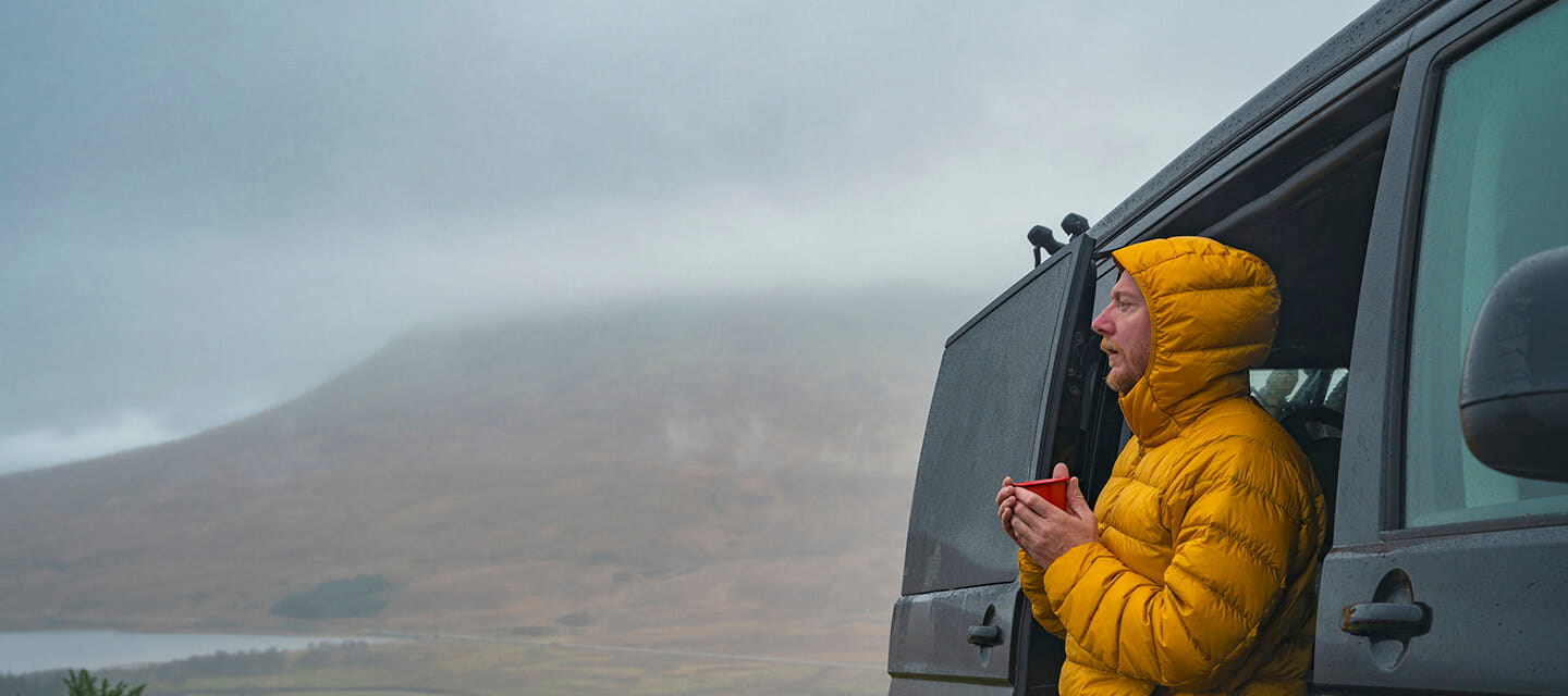 A mature man takes in the view of the Scottish Highlands on a rainy day from his campervan. He wears his hooded puffer coat and warms himself up with a cup of tea.