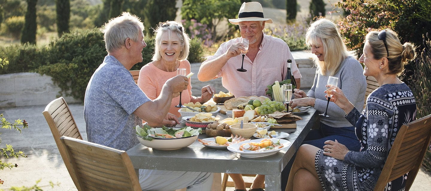 A group of mature friends are sitting around an outdoor dining table, eating and drinking in Tuscany, Italy.