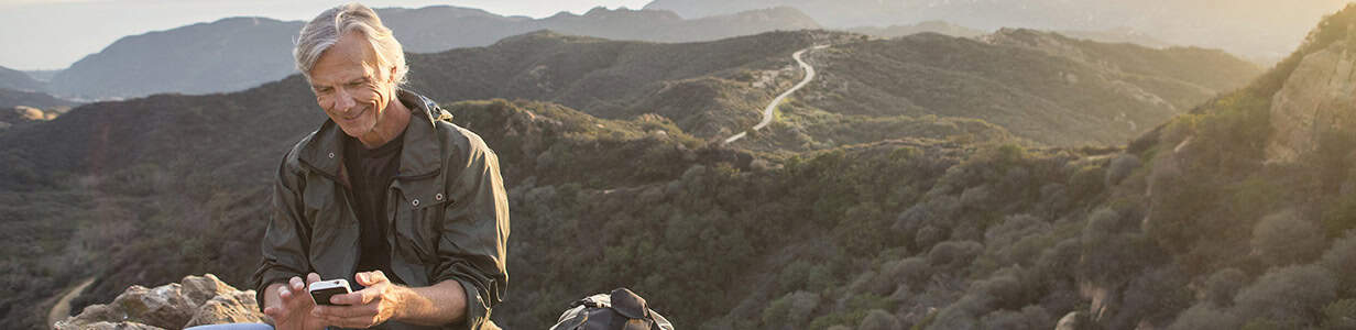 A mature man smiling at his phone with a majestic forest sprawling out in the background.