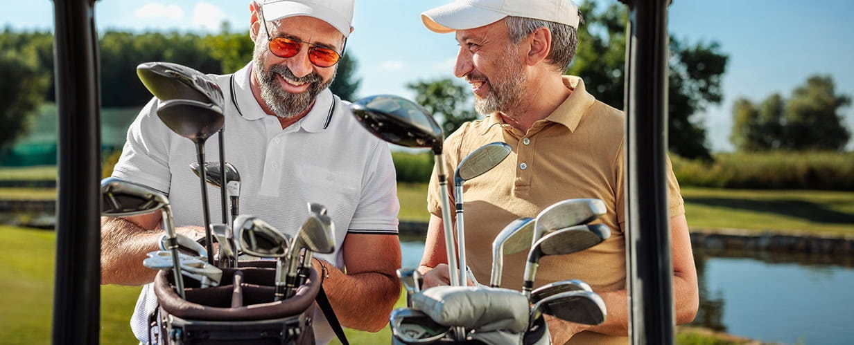 Two older golf players taking golf clubs from a golf cart while talking and smiling on the field