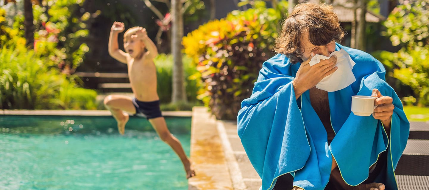 Poorly man caught a cold on holiday, sits sad at the pool drinking tea and blows his nose into a napkin. His grandson is healthy and swimming in the pool.