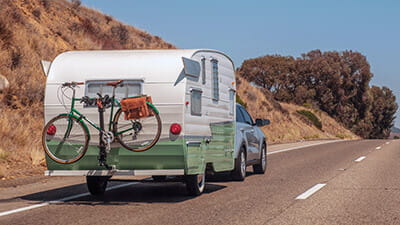 A retro caravan with a green bicycle on the back traveling on a country road.