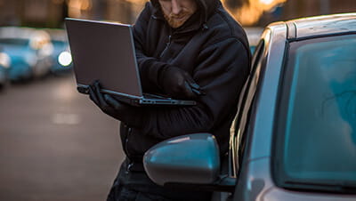 A keyless auto criminal dressed in black with tools of the trade hacking into the security of a car with a laptop in the early morning