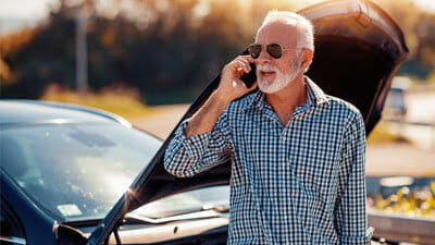 A senior man calling for assistance on his mobile phone next to a broken down car