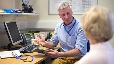 A male doctor sits face to face with a mature woman in the doctors office.