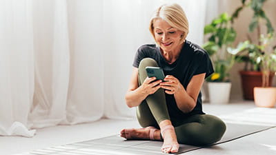 Mature woman texting on her smartphone after doing her daily workout at home on yoga mat.