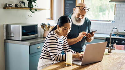 A mature couple using a laptop and phone in the kitchen at home.