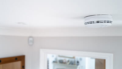 Shallow focus of a hard wired smoke detector seen in the downstairs hall, fixed to the ceiling. An open door leading to a living room can be seen.