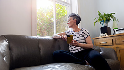 A mature female adult sitting on a leather sofa. She has her phone in her hands whilst looking out the window.