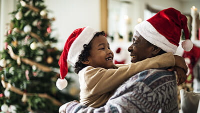 A grandfather holding his grandson in front of a Christmas tree, both wearing Christmas hats