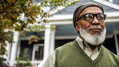 A stylish mature man with a grey beard standing in front of his house