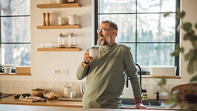 A content man smiling to himself in his kitchen while drinking coffee.