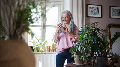 Fashionable senior woman standing by desk and text messaging at home