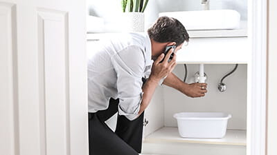 A man calling a plumber in front of water leaking from a sink pipe