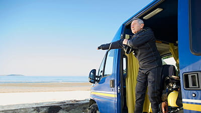 A senior diver puts on a wetsuit in his camper van parked beside the beach.