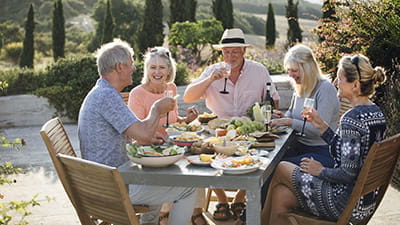A group of mature friends are sitting around an outdoor dining table, eating and drinking in Tuscany, Italy.