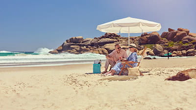 A mature couple sitting together under a parasol on a sandy beach.