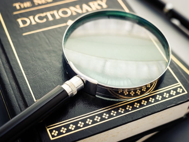 A magnifying glass resting on a copy of an English dictionary | Getty/patpitchaya