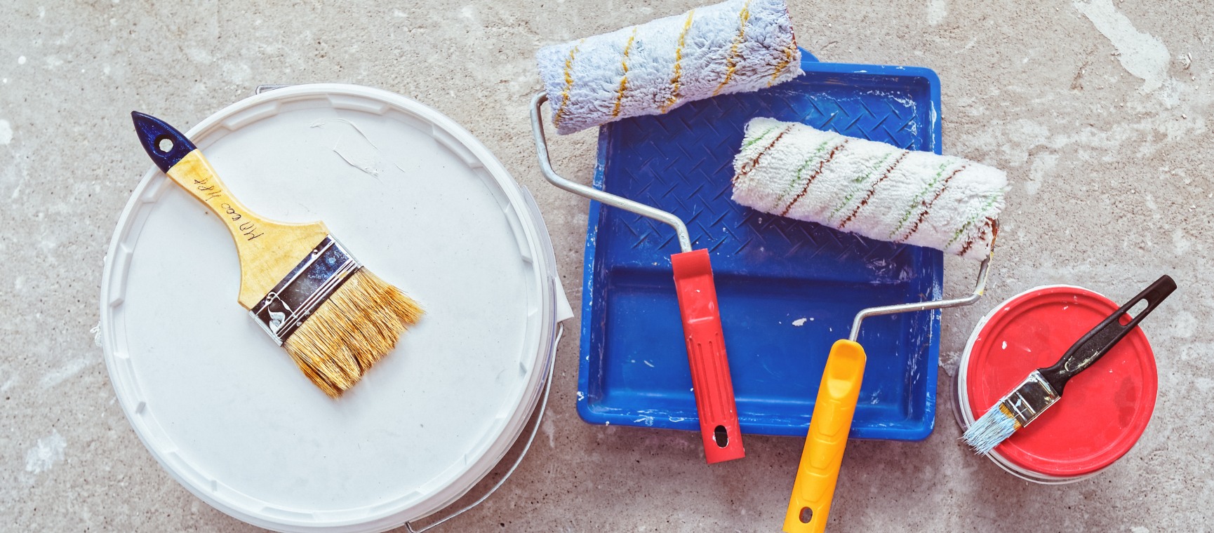 A styled image of a clean paint roller. orange paint pot and orange painting tray, against a bright blue background