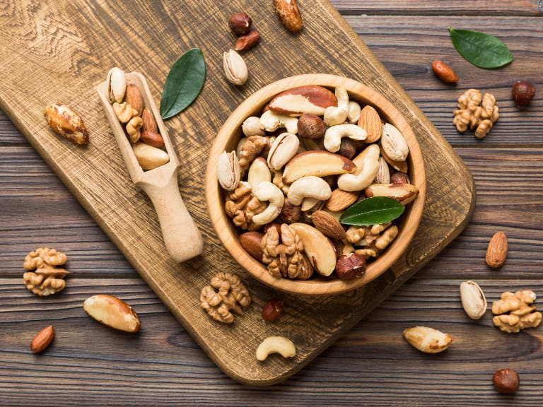 A bowl of mixed nuts displayed on a wooden board with a wooden scoop and scattered nuts