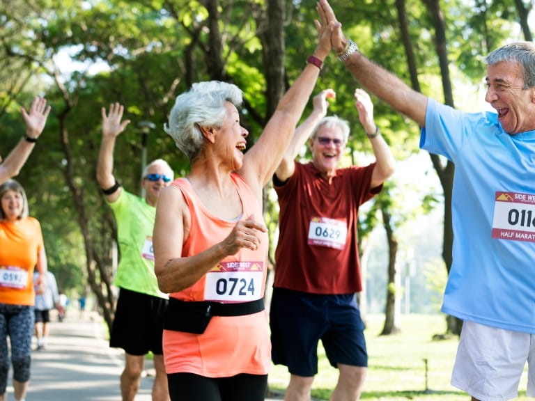 A group of people running a race wearing race numbers; at the front, a mature woman is high fiving a man