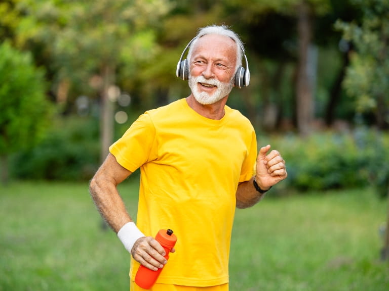 An older man with a white beard wears a bright yellow t-shirt and headphones while running outdoors, holding an orange water bottle