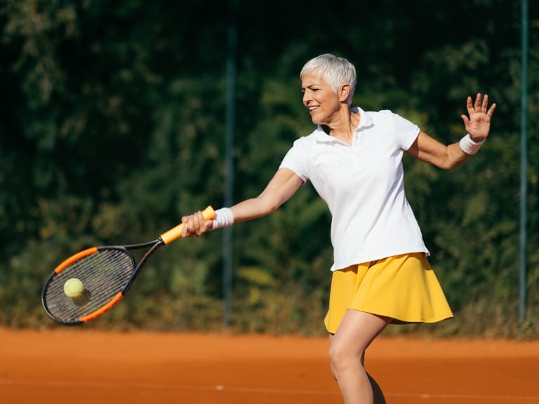 Woman with cropped hair wears a white t-shirt and yellow tennis skirt while playing tennis on an outdoor court