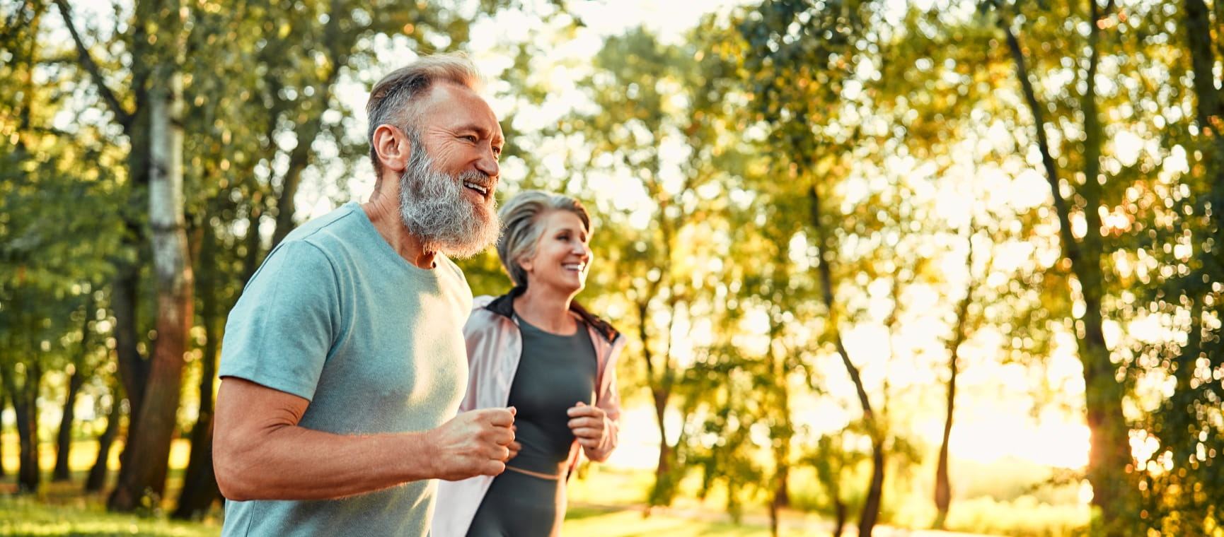 A bearded man and smiling woman enjoying a run outdoors with autumnal trees behind them