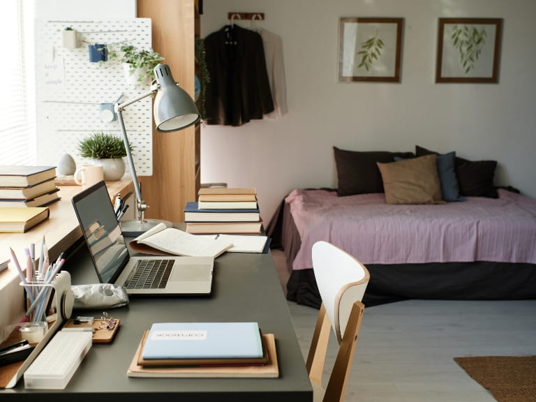 A home office turned spare bedroom with a desk, laptop, lamp and chair, and single bed in the background