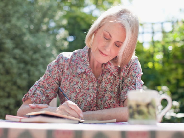 An older woman wearing a floral shirt while writing in a journal outdoors with a cup of tea