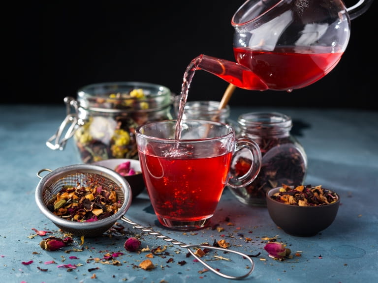 Red herbal tea being poured from a glass teapot into a cup, surrounded by a display of herbs