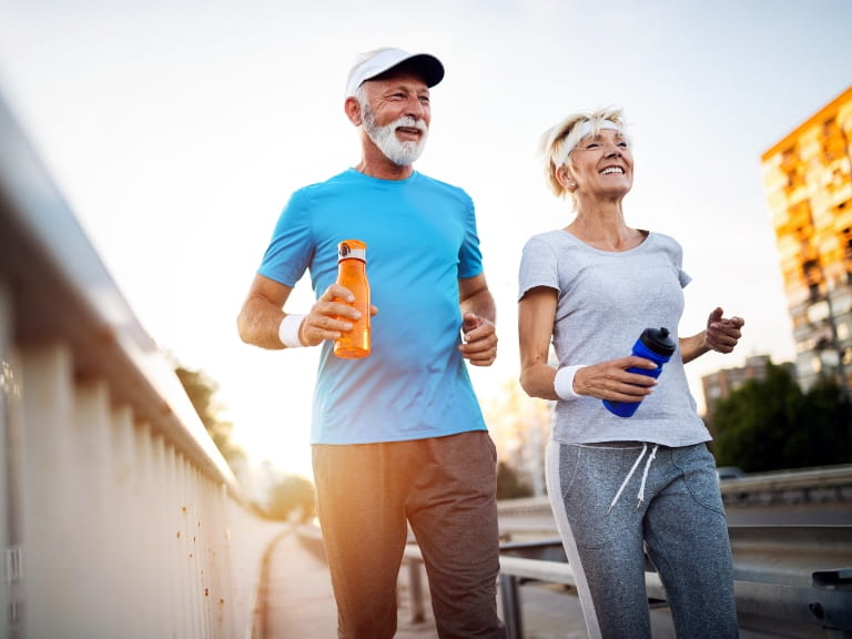 Mature couple running across a bridge in a city in sports attire while carrying water bottles