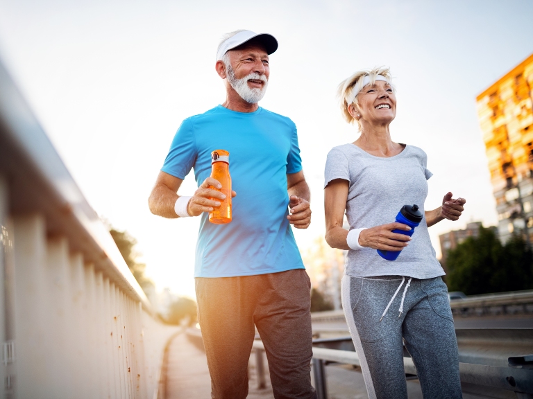 Mature couple running across a bridge in a city in sports attire while carrying water bottles