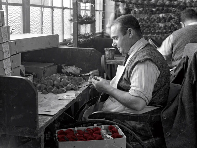 A mostly black-and-white photograph of a male worker in the Poppy Factory assembling poppies for Remembrance Sunday; the poppies are red