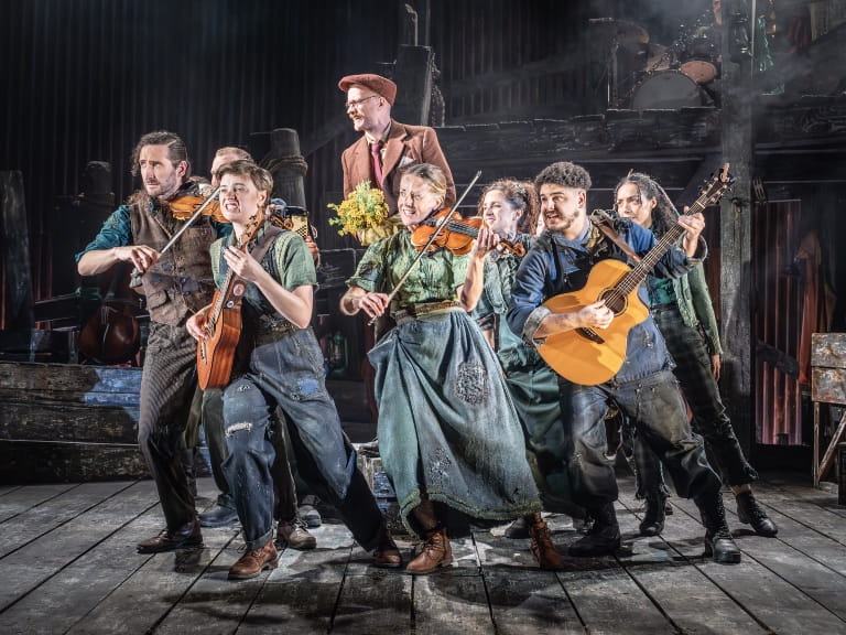 An ensemble of actors playing instruments and singing during a stage production of The Curious Case of Benjamin Button