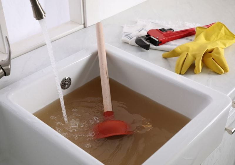 A white kitchen sink full of dirty water with a plunger half submerged, with a wrench and rubber gloves on the side