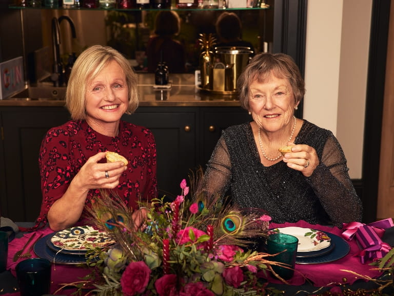 Two women eating mince pies at a table adorned with Christmas decorations