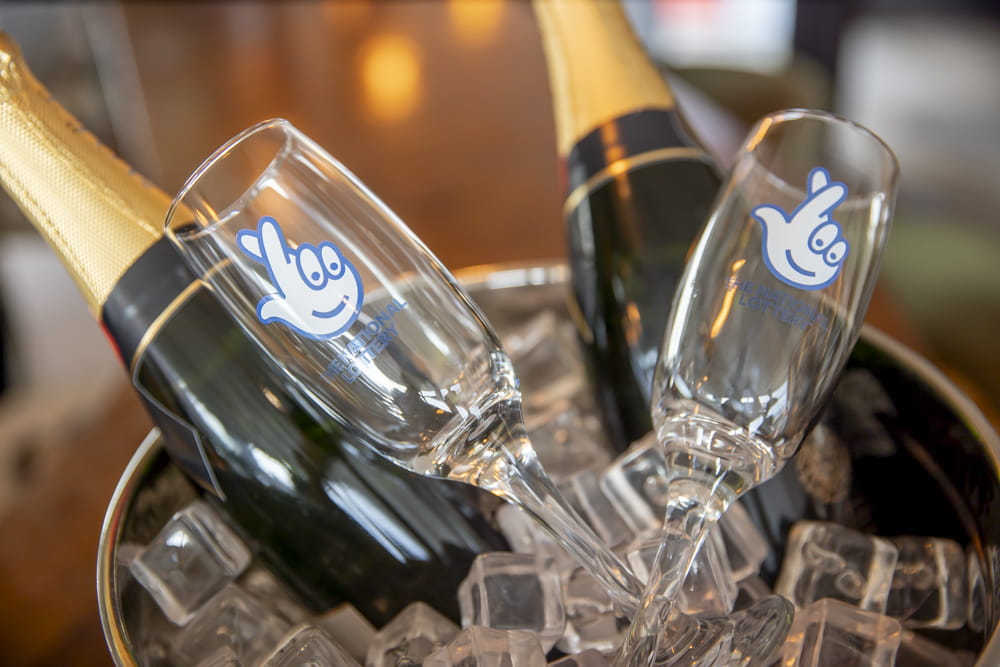 Two champagne bottles in an ice bucket with flute glasses with the National Lottery logo