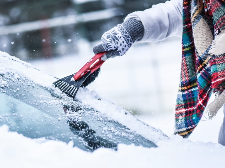 Someone wearing gloves and a tartan scarf scraping snow off a car windscreen