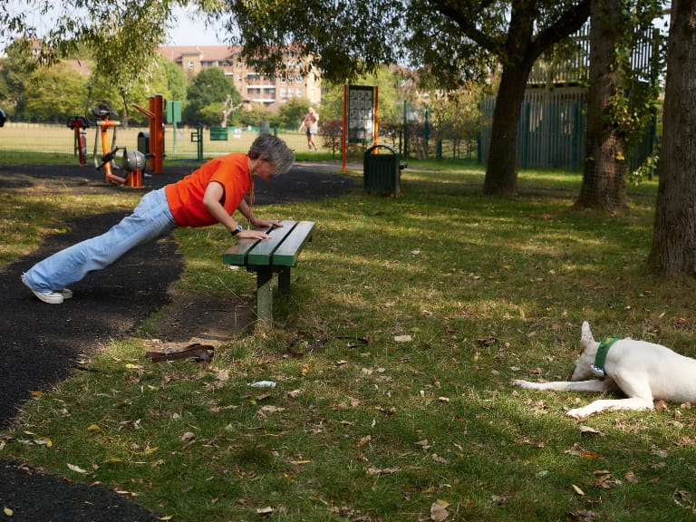 A woman doing a bench press-up in a park with her dog nearby
