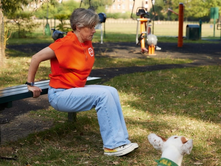 A woman doing a tricep dip on a park bench with her dog nearby