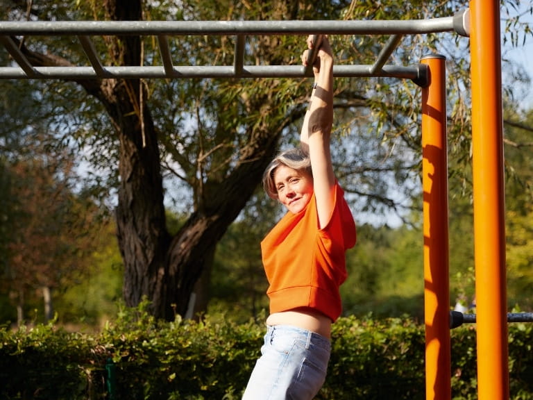 A woman using bars to do a dead hang in the park with her dog nearby