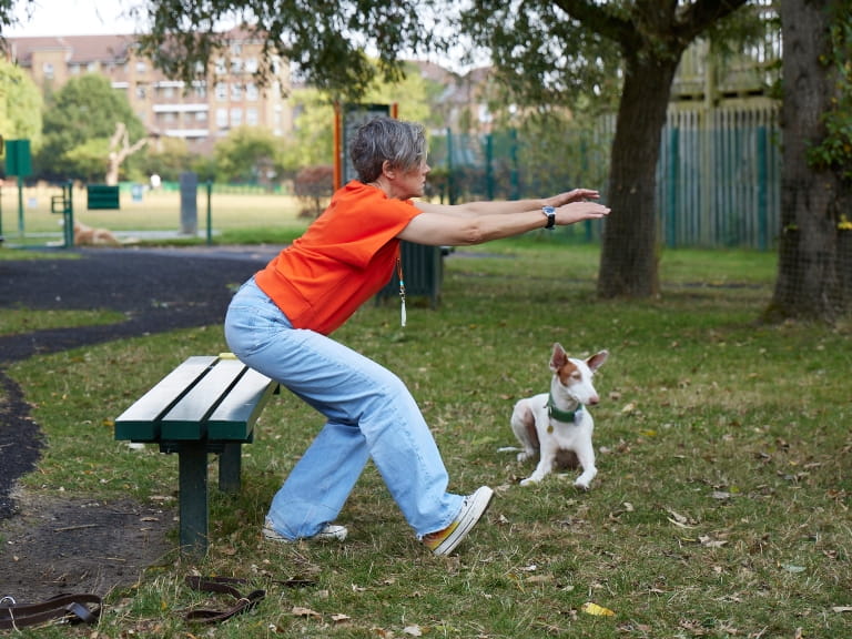 A woman doing a one-leg squat in front of a park bench with her dog nearby