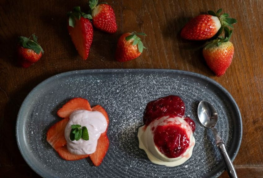 A grey plate with sliced strawberries and panna cotta on top and next to it panna cotta with red berry sauce